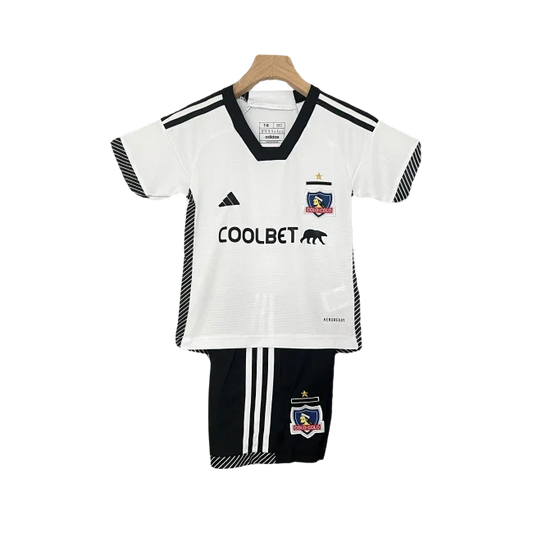 Kit infantil Colo Colo I 24/25, com camisa branca gola V preta, logos Adidas e Coolbet pretos, escudo. Shorts pretos com listras brancas.