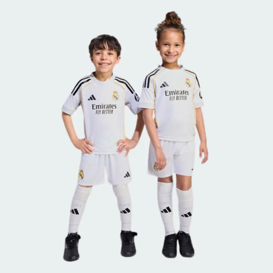 Dois jovens sorrindo, um menino e uma menina, vestindo o kit infantil Real Madrid I 25/26 branco com detalhes em preto e dourado.
