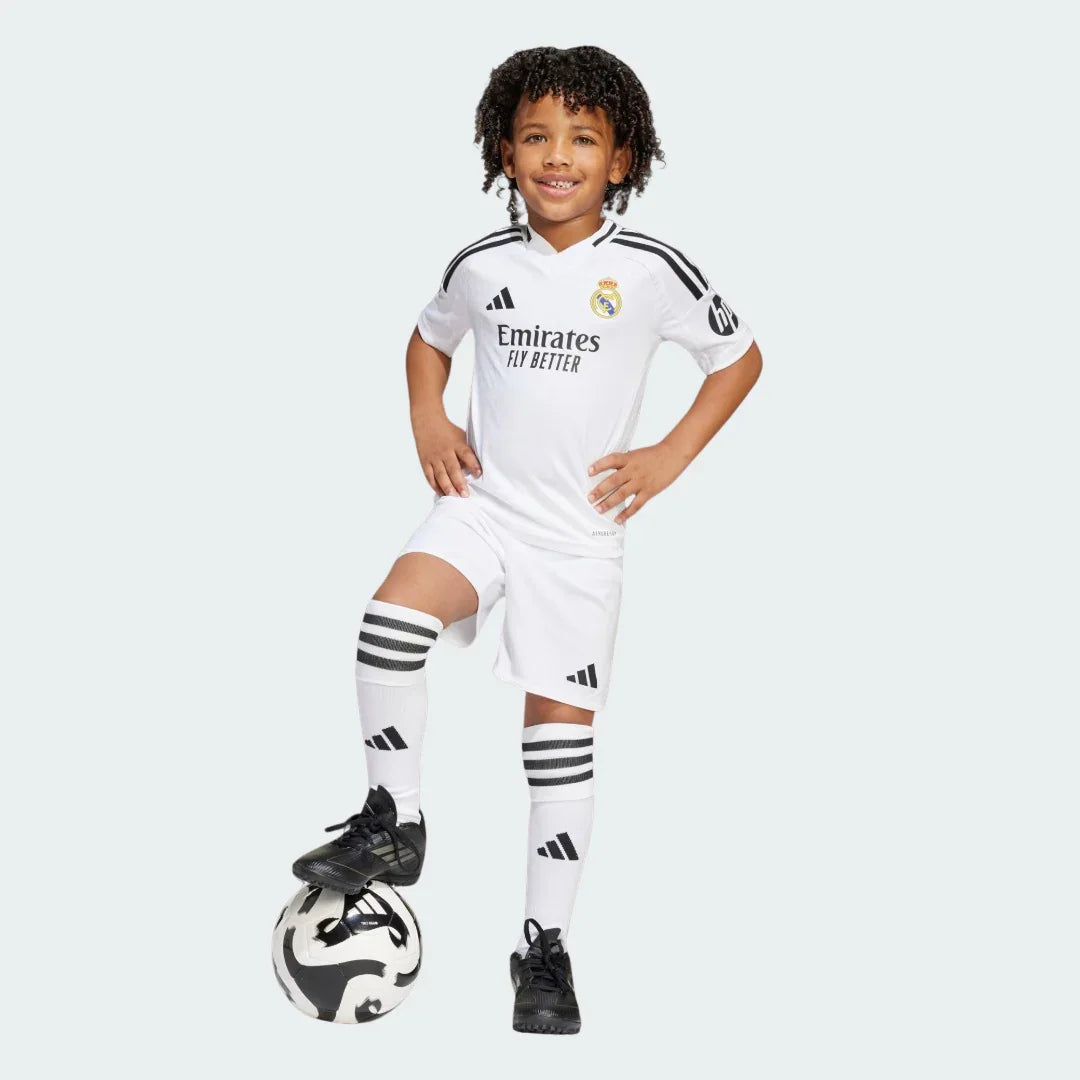 Criança sorridente veste kit infantil Real Madrid branco com detalhes pretos e segura bola de futebol.