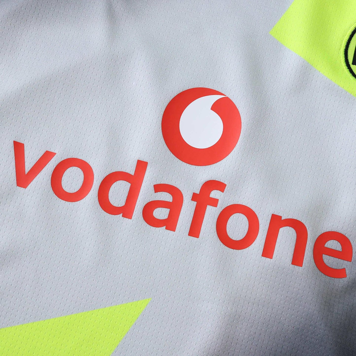 Detalhe da camisa infantil cinza-clara com o logotipo da Vodafone em laranja-avermelhado e detalhes em amarelo neon.