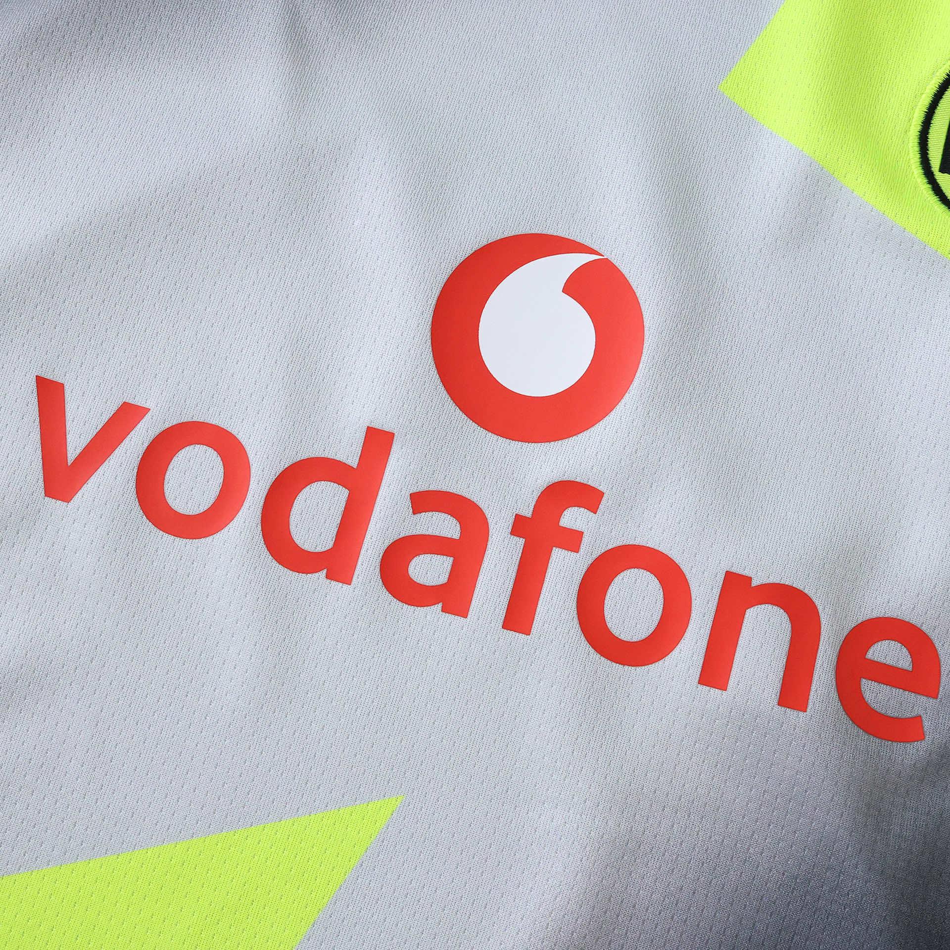 Detalhe da camisa infantil cinza-clara com o logotipo da Vodafone em laranja-avermelhado e detalhes em amarelo neon.