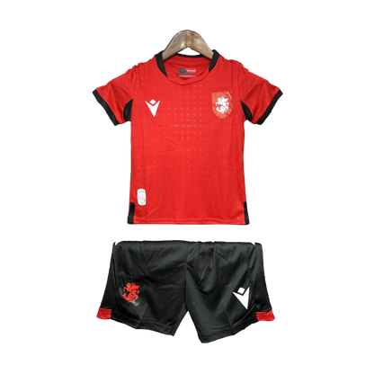 Kit de futebol infantil da Georgia 24/25 com camisa vermelha e shorts pretos, exibido em cabide contra fundo preto.