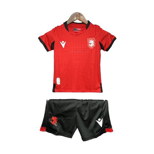 Kit de futebol infantil da Georgia 24/25 com camisa vermelha e shorts pretos, exibido em cabide contra fundo preto.