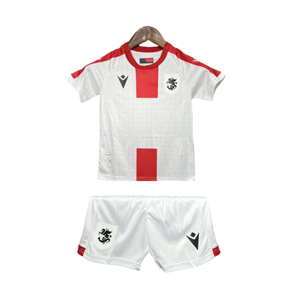 Kit infantil Georgia 24/25, com camisa e shorts brancos. Camisa com faixas vermelhas, escudo preto e logo Macron preto. Shorts brancos com escudo e logo Macron pretos.