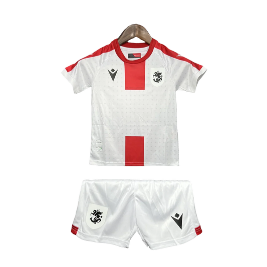 Kit infantil Georgia 24/25, com camisa e shorts brancos. Camisa com faixas vermelhas, escudo preto e logo Macron preto. Shorts brancos com escudo e logo Macron pretos.
