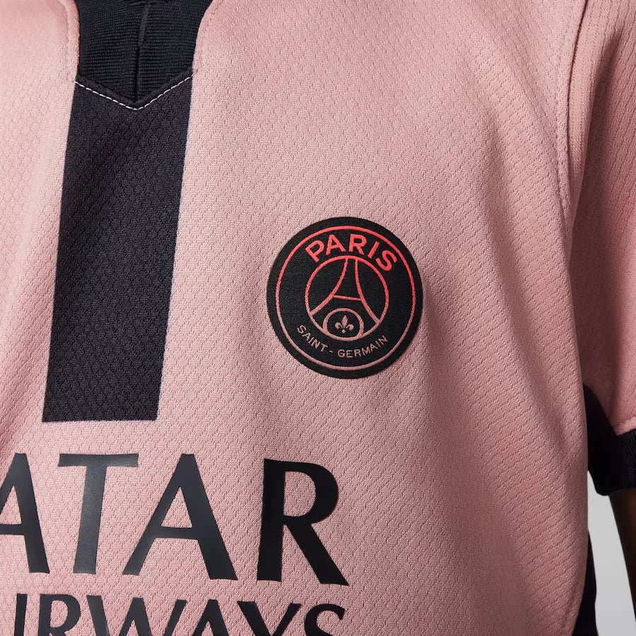 Close-up da camisa rosa do PSG, mostrando o escudo redondo do clube e o logo do patrocinador, com detalhes em preto na gola e faixa vertical.