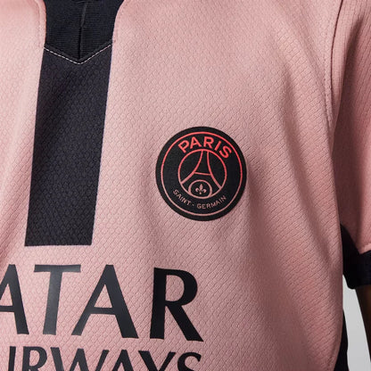 Close-up da camisa rosa do PSG, mostrando o escudo redondo do clube e o logo do patrocinador, com detalhes em preto na gola e faixa vertical.