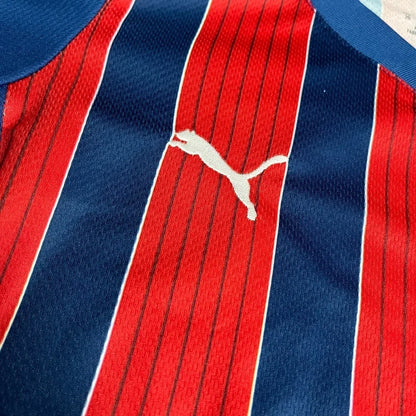 Camisa de futebol infantil listrada verticalmente em vermelho e azul escuro, com o logo branco da Puma no peito.
