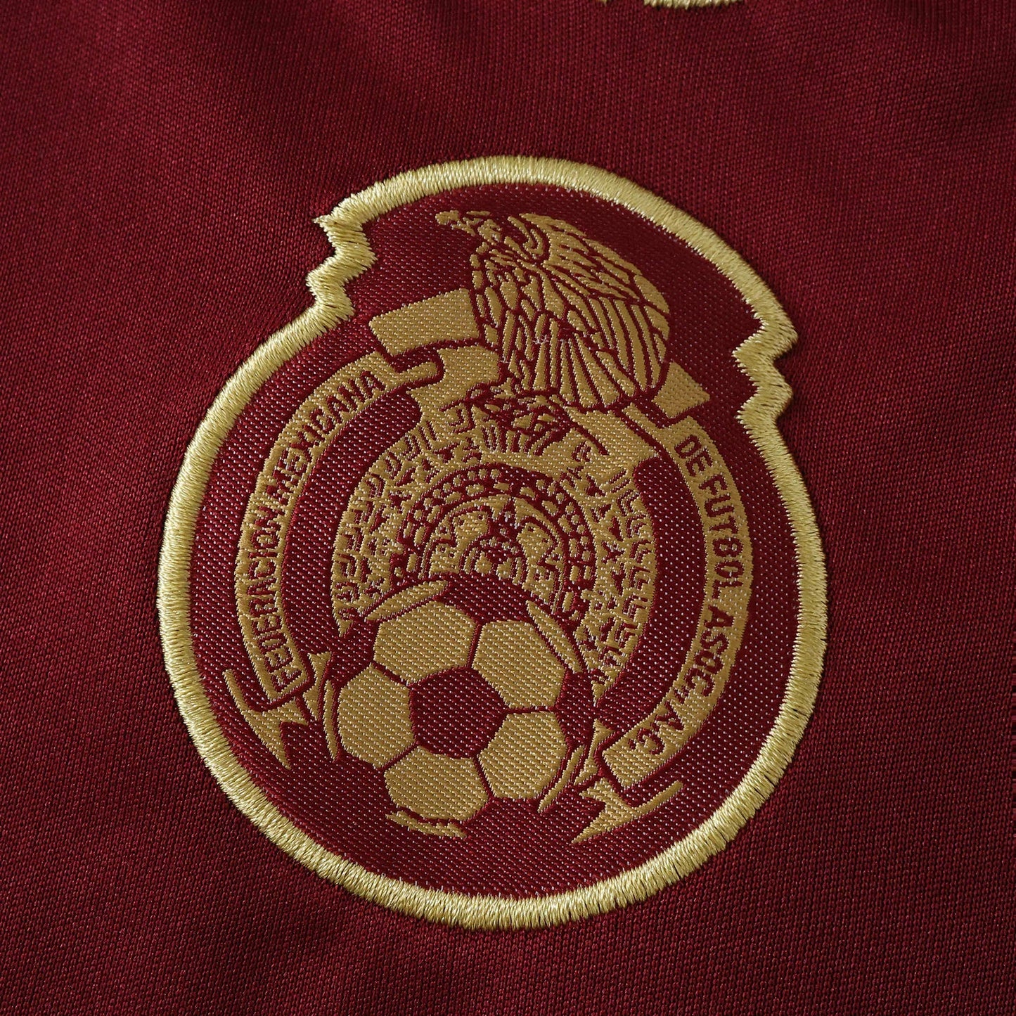 Detalhe de um escudo bordado em dourado em tecido de camisa na cor vinho, com uma águia, bola de futebol e inscrições.