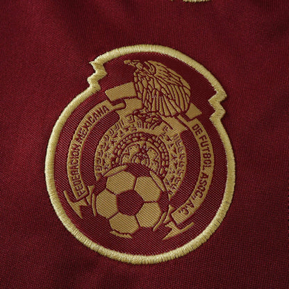 Detalhe de um escudo bordado em dourado em tecido de camisa na cor vinho, com uma águia, bola de futebol e inscrições.