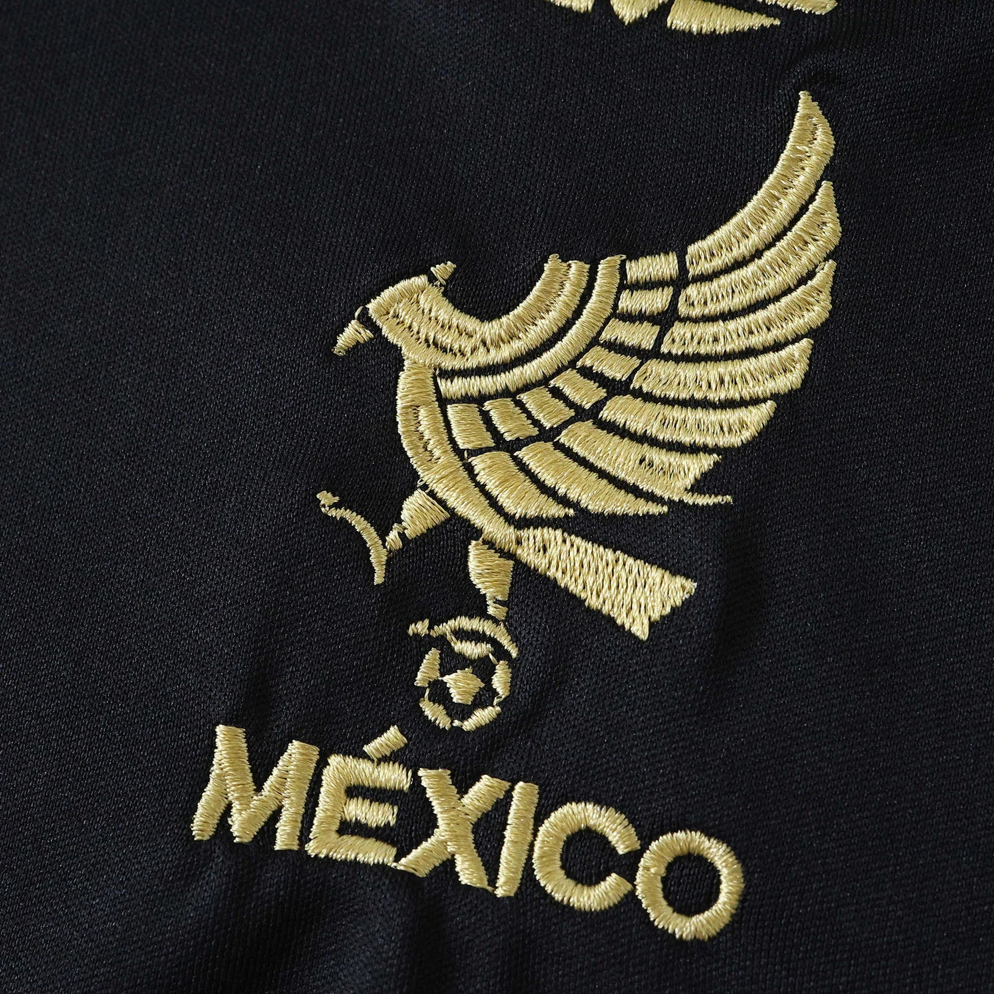 Close-up da camisa de futebol preta com escudo bordado dourado de águia segurando uma bola e a palavra MÉXICO em dourado.