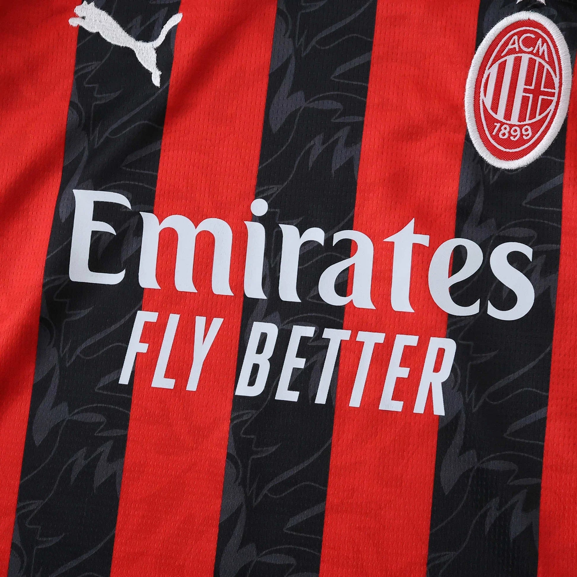 Detalhe da camisa infantil AC Milan. Listras verticais vermelhas e pretas com padrão, logo Puma, escudo ACM e patrocínio Emirates.