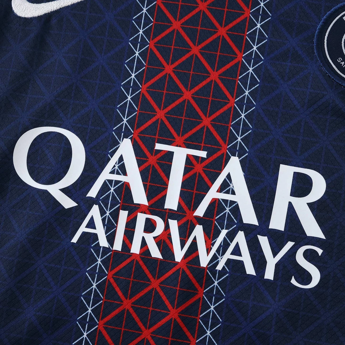 Detalhe de camisa de futebol infantil azul-marinho com padrão geométrico, patrocínio 'QATAR AIRWAYS' em branco, logo Nike e escudo circular com 'SAI'.