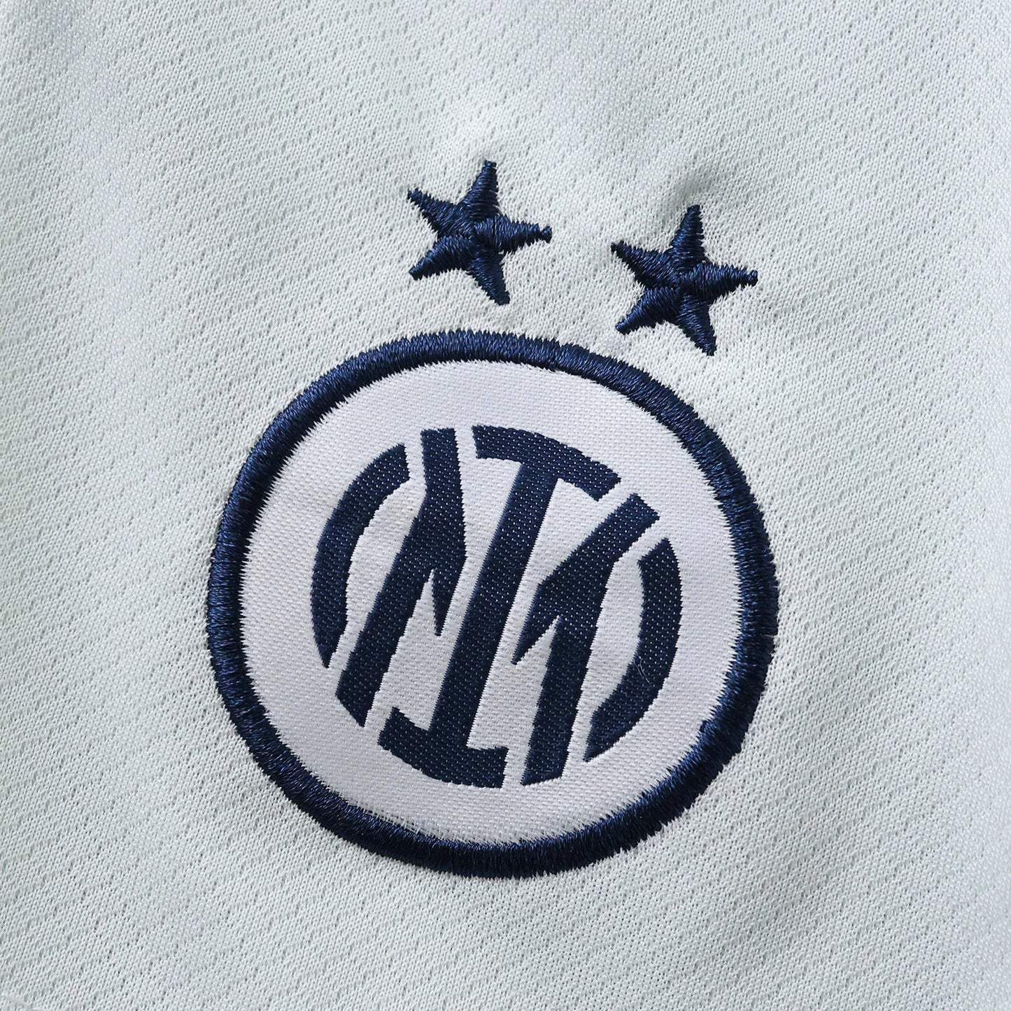 Close-up do distintivo bordado da Inter Milan em azul escuro e branco, com duas estrelas azuis, sobre um tecido azul claro.