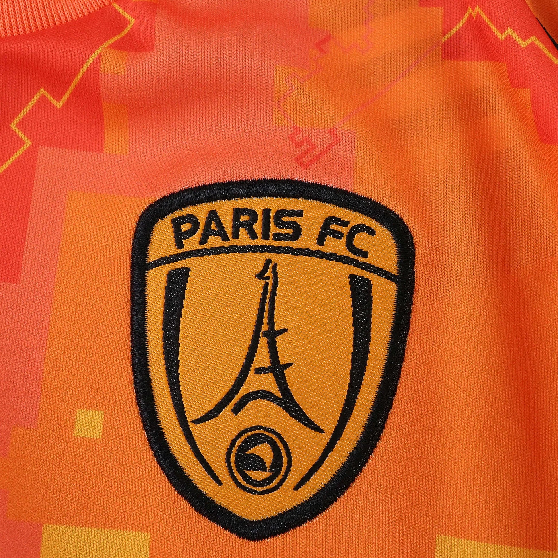 Camisa laranja Paris FC, detalhe do escudo bordado em preto com Torre Eiffel e fundo laranja claro.