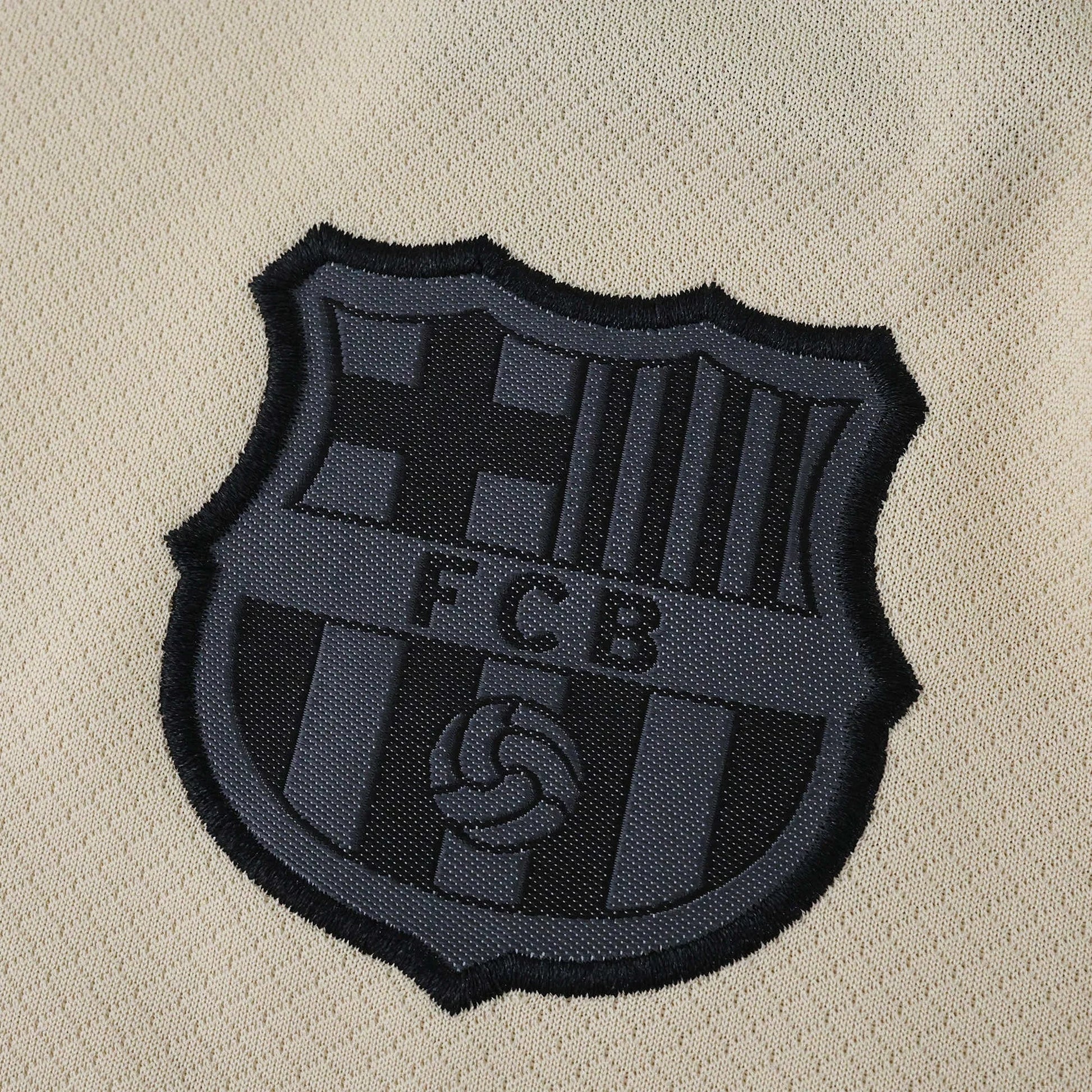 Close-up do escudo escuro em relevo com as letras FCB em uma camisa infantil de futebol na cor bege claro.