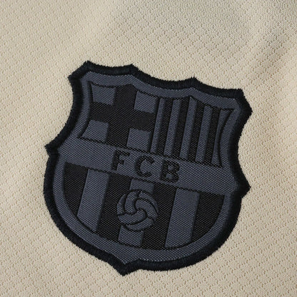 Close-up do escudo escuro em relevo com as letras FCB em uma camisa infantil de futebol na cor bege claro.