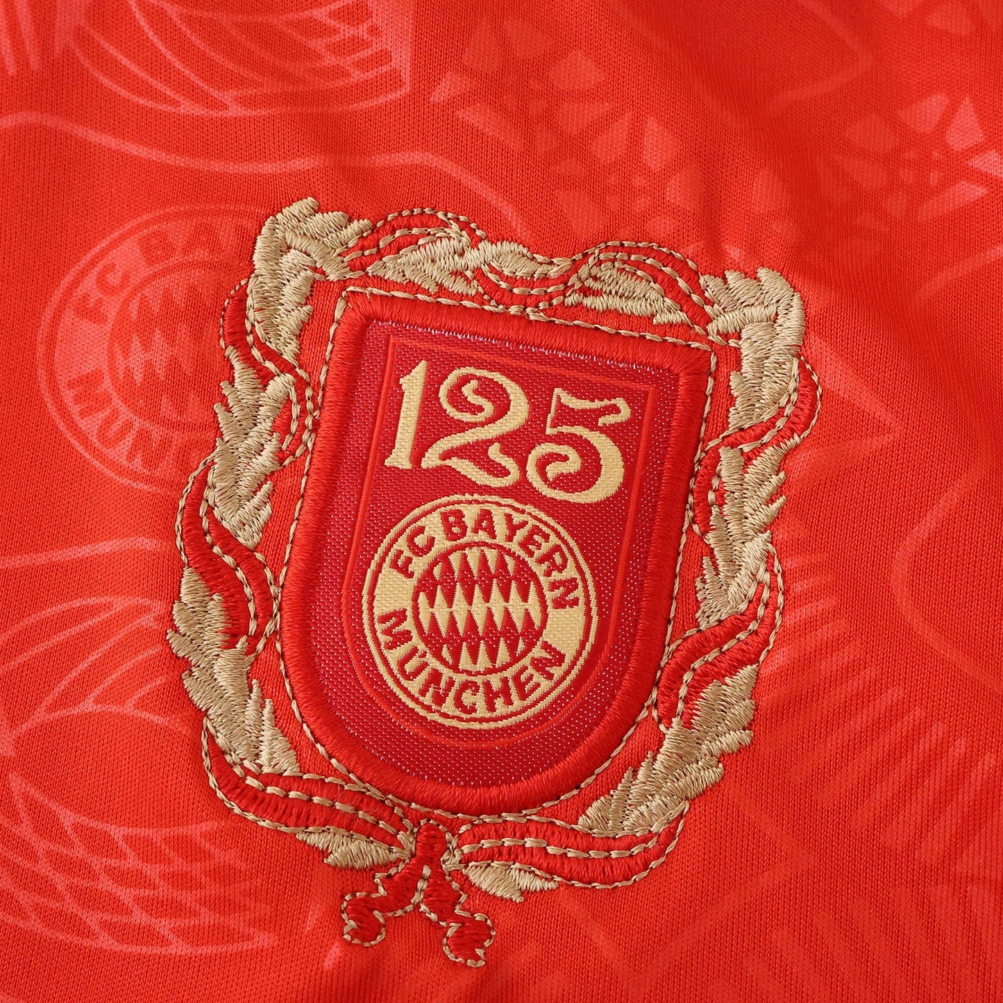 Escudo comemorativo de 125 anos do FC Bayern München bordado em dourado e vermelho sobre tecido de camisa vermelha.