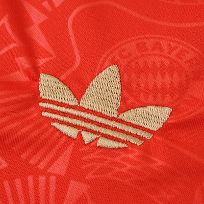 Close-up de camisa vermelha. Logo Adidas Originals bordado em bege no centro. Padrão em relevo com letras e detalhes gráficos no tecido.
