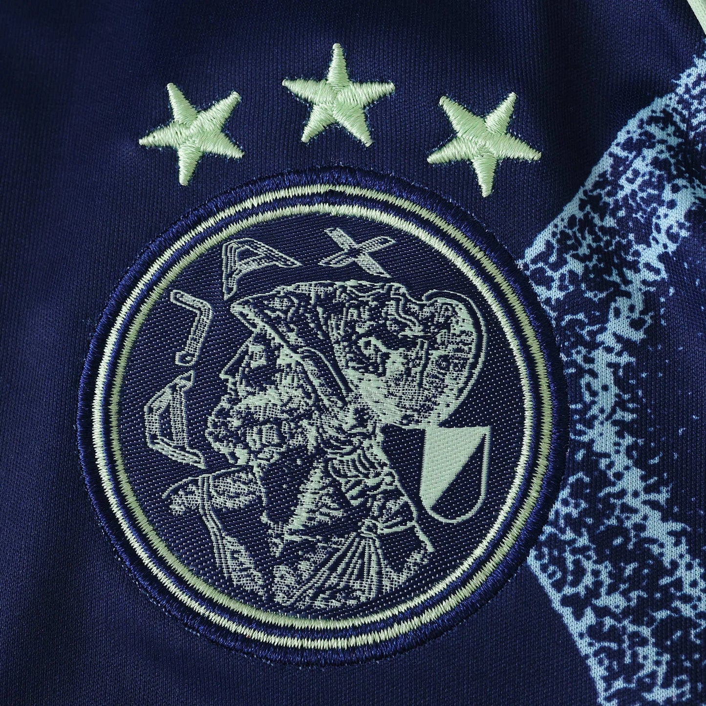 Emblema bordado do Ajax com três estrelas em verde claro sobre tecido azul marinho, detalhe lateral azul com manchas.
