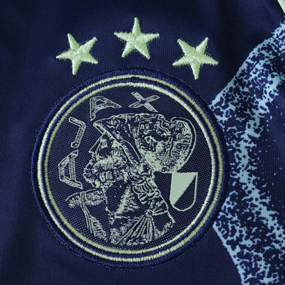 Emblema bordado do Ajax com três estrelas em verde claro sobre tecido azul marinho, detalhe lateral azul com manchas.