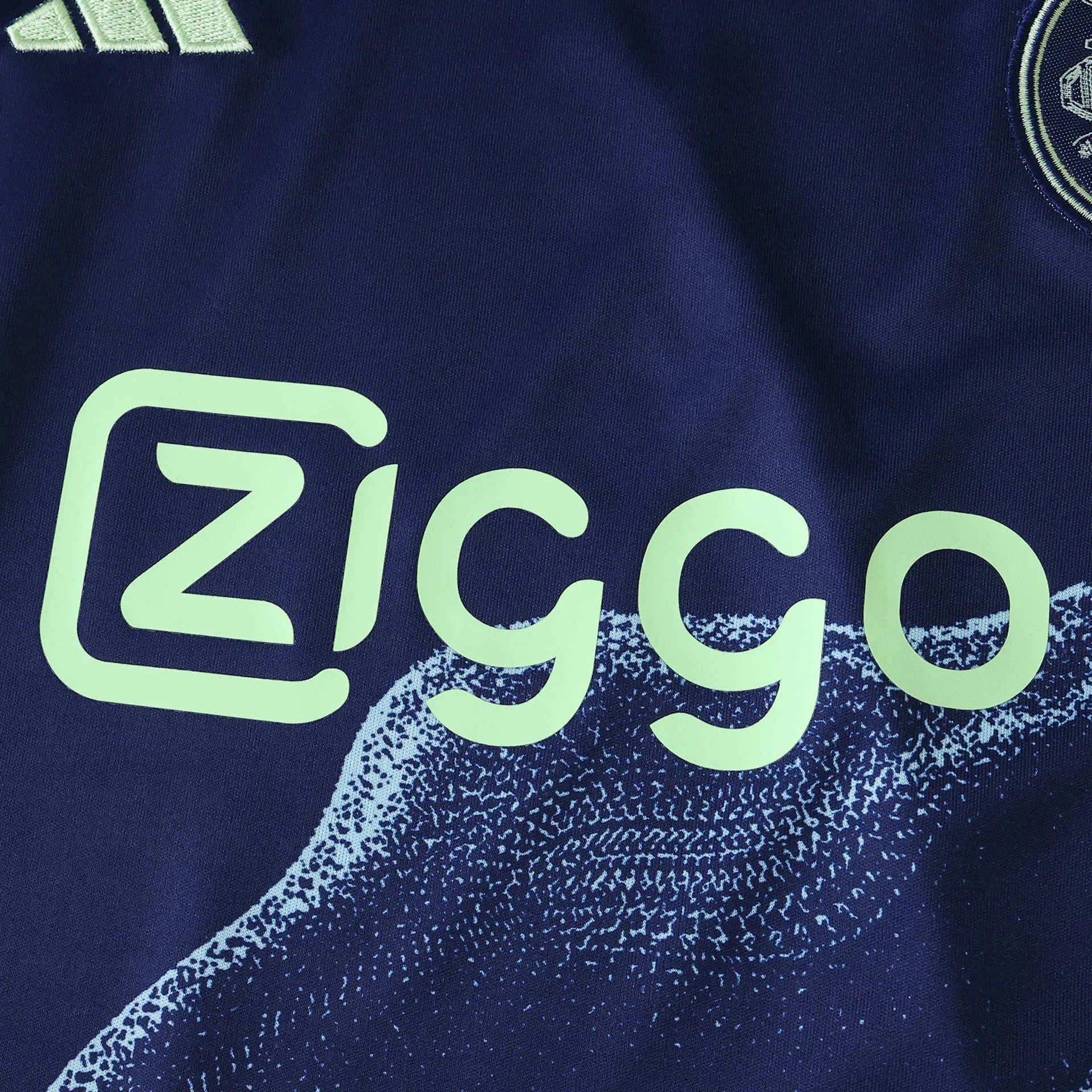 Close-up da camisa azul-marinho do AJAX, destacando o logo Ziggo verde-limão e detalhes do escudo e Adidas.