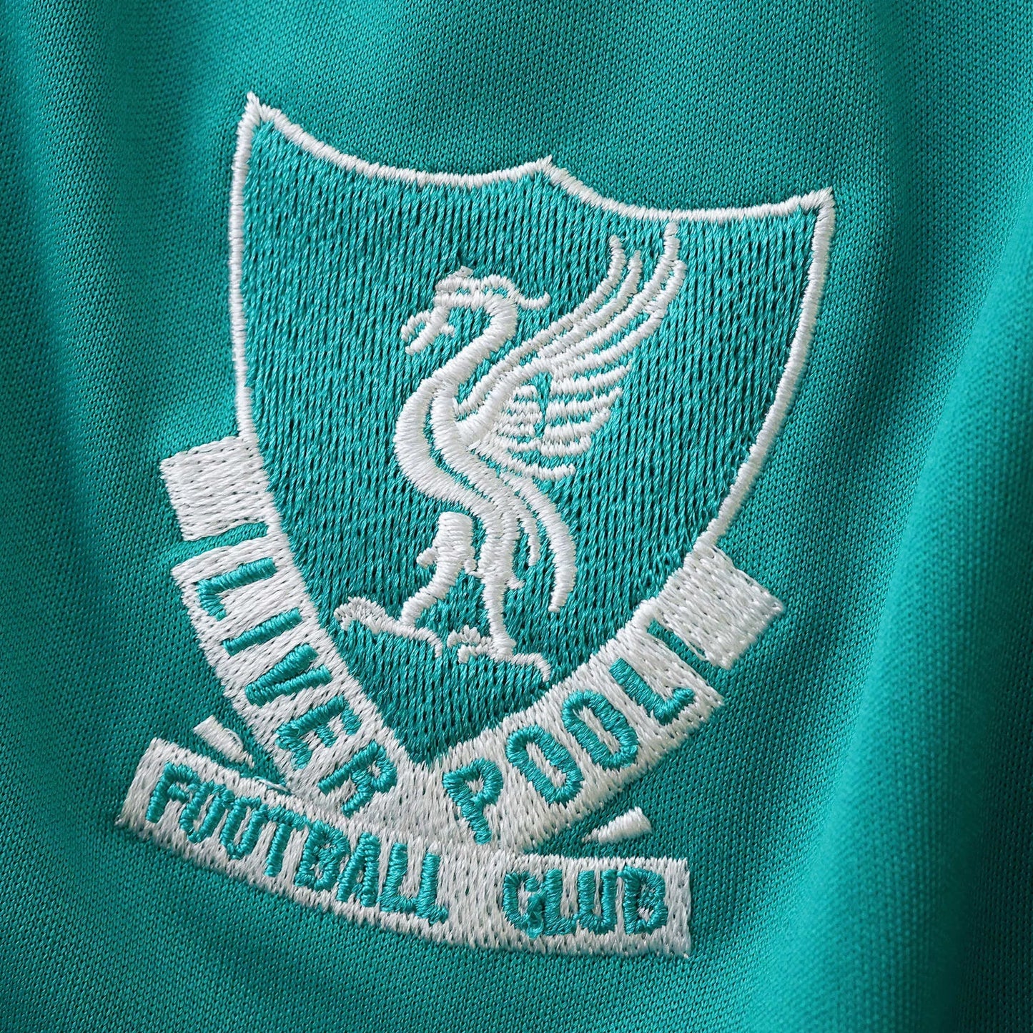 Close-up do escudo bordado do Liverpool Football Club em uma camisa infantil na cor teal. O escudo apresenta o Liver Bird em branco.