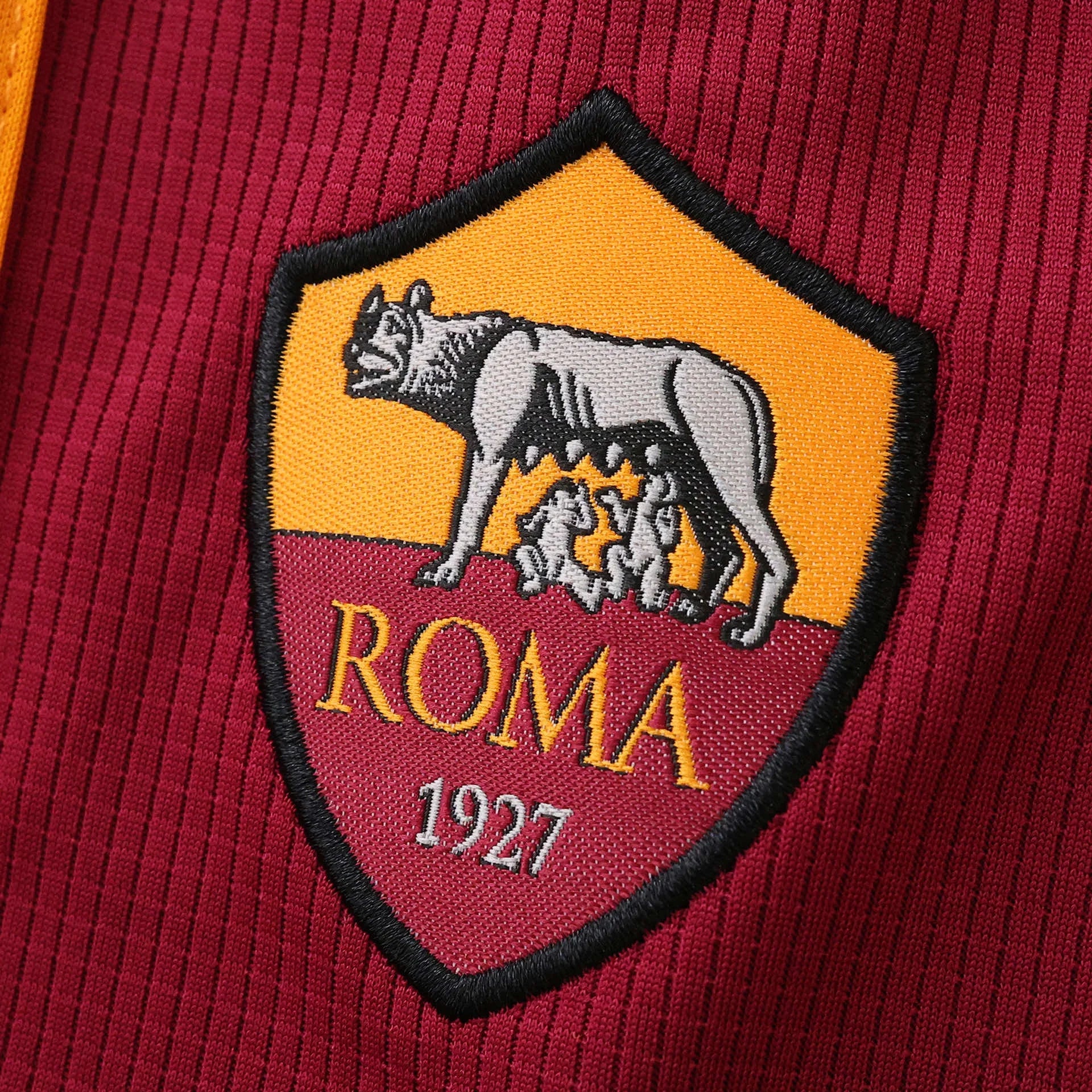 Detalhe do escudo bordado da camisa da AS Roma, com a loba e os números 'ROMA 1927' em fundo laranja e bordô, sobre tecido canelado.