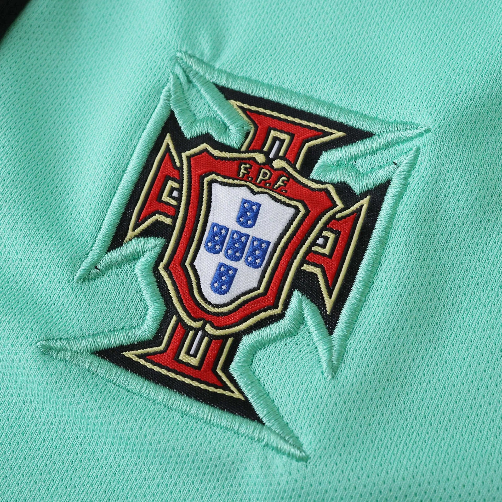 Detalhe do escudo bordado da seleção de Portugal em camisa verde-água, com emblema F.P.F. em vermelho, branco, azul e dourado.