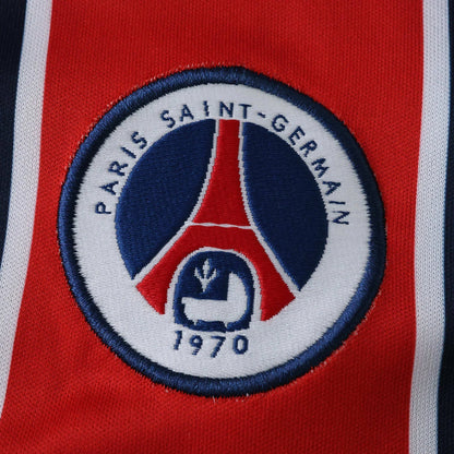 Close-up do distintivo bordado do Paris Saint-Germain em uma camisa vermelha, com listras laterais brancas e azul-escuras.