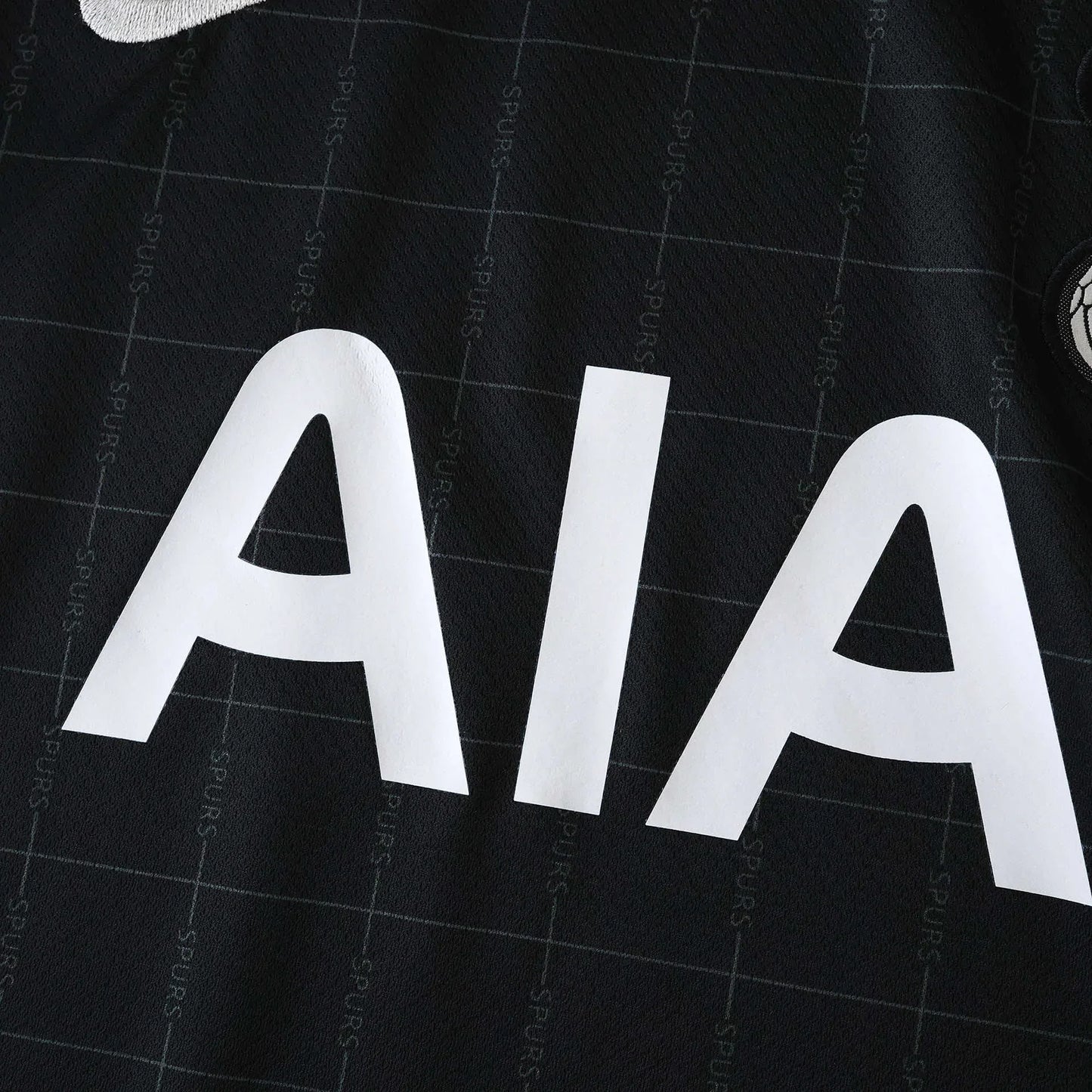 Close-up da camisa preta do Tottenham Hotspur com o patrocínio branco AIA e padrão sutil de SPURS.
