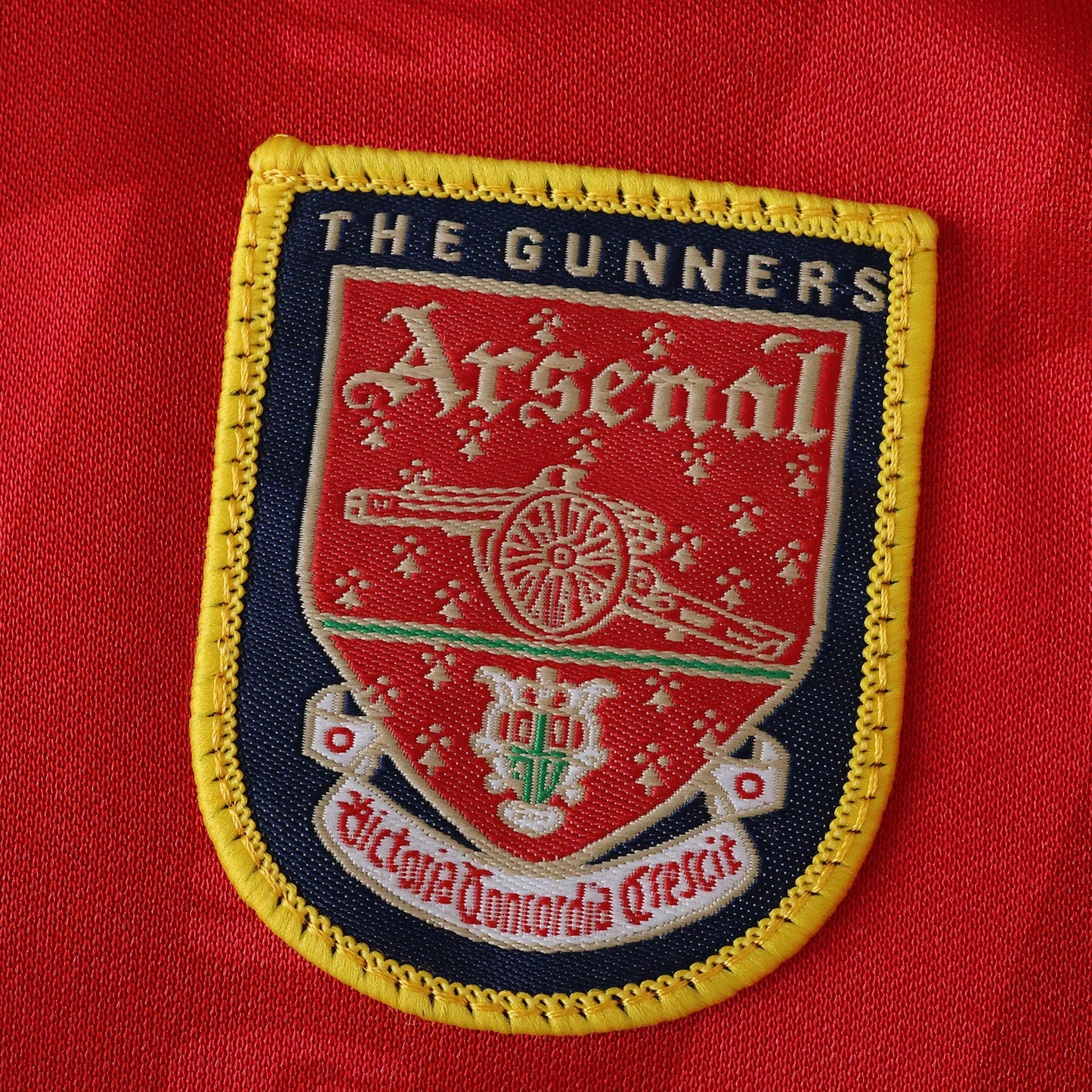 Emblema bordado do Arsenal em camisa vermelha. Detalha o escudo com canhão, 'THE GUNNERS', 'Arsenal' e 'Victoria Concordia Crescit'.