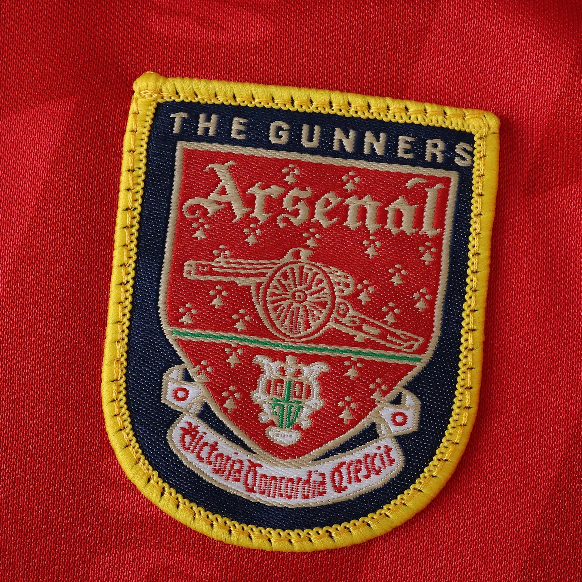 Emblema bordado do Arsenal em camisa vermelha. Detalha o escudo com canhão, 'THE GUNNERS', 'Arsenal' e 'Victoria Concordia Crescit'.
