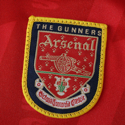 Emblema bordado do Arsenal em camisa vermelha. Detalha o escudo com canhão, 'THE GUNNERS', 'Arsenal' e 'Victoria Concordia Crescit'.