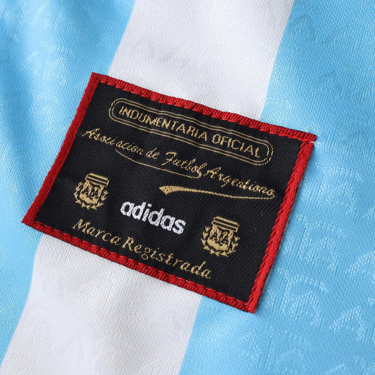 Close-up da etiqueta oficial preta da AFA e Adidas com bordas vermelhas, sobre tecido de camisa listrada azul claro e branca.