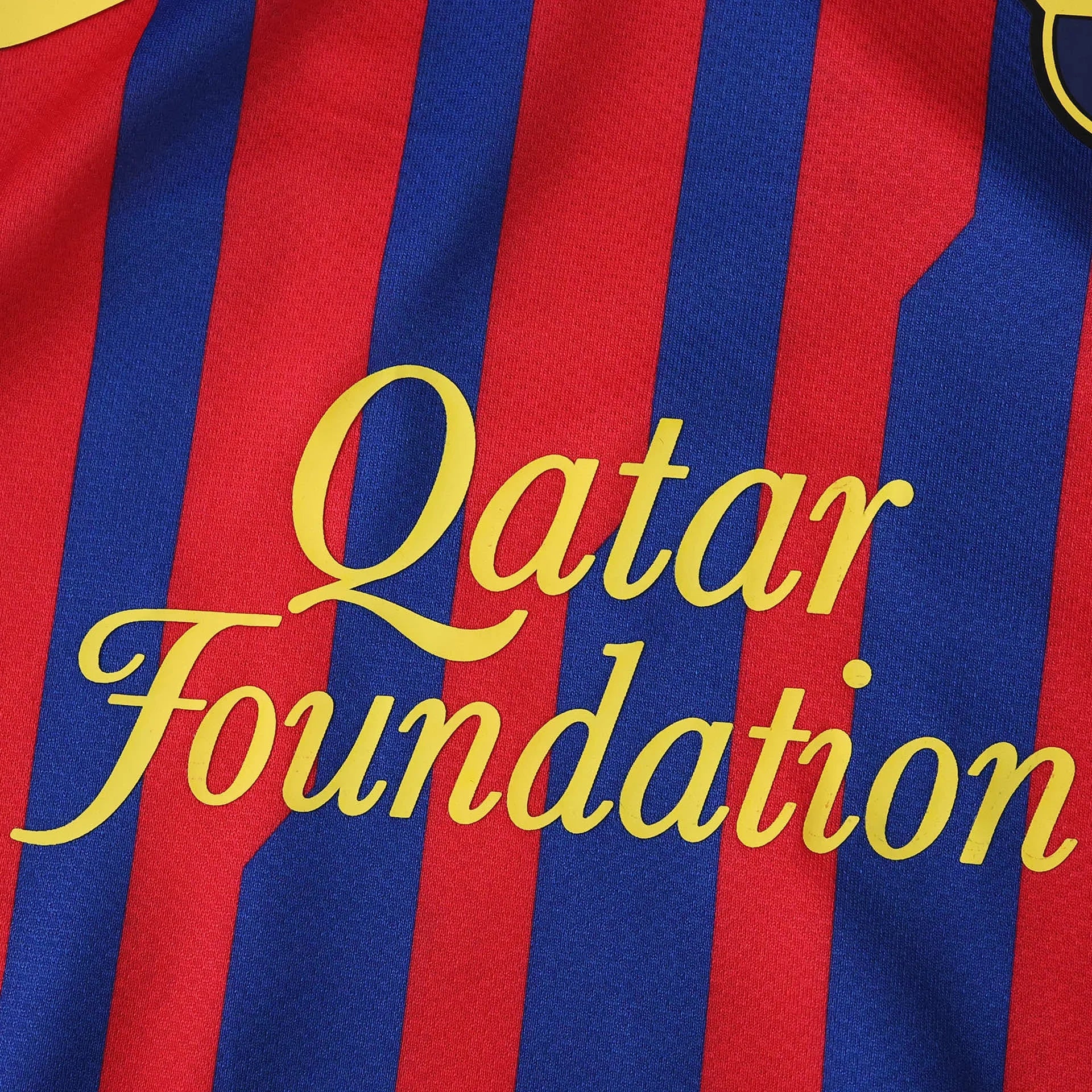 Camisa de futebol infantil com listras verticais azuis e vermelhas, e patrocínio 'Qatar Foundation' em letras amarelas.