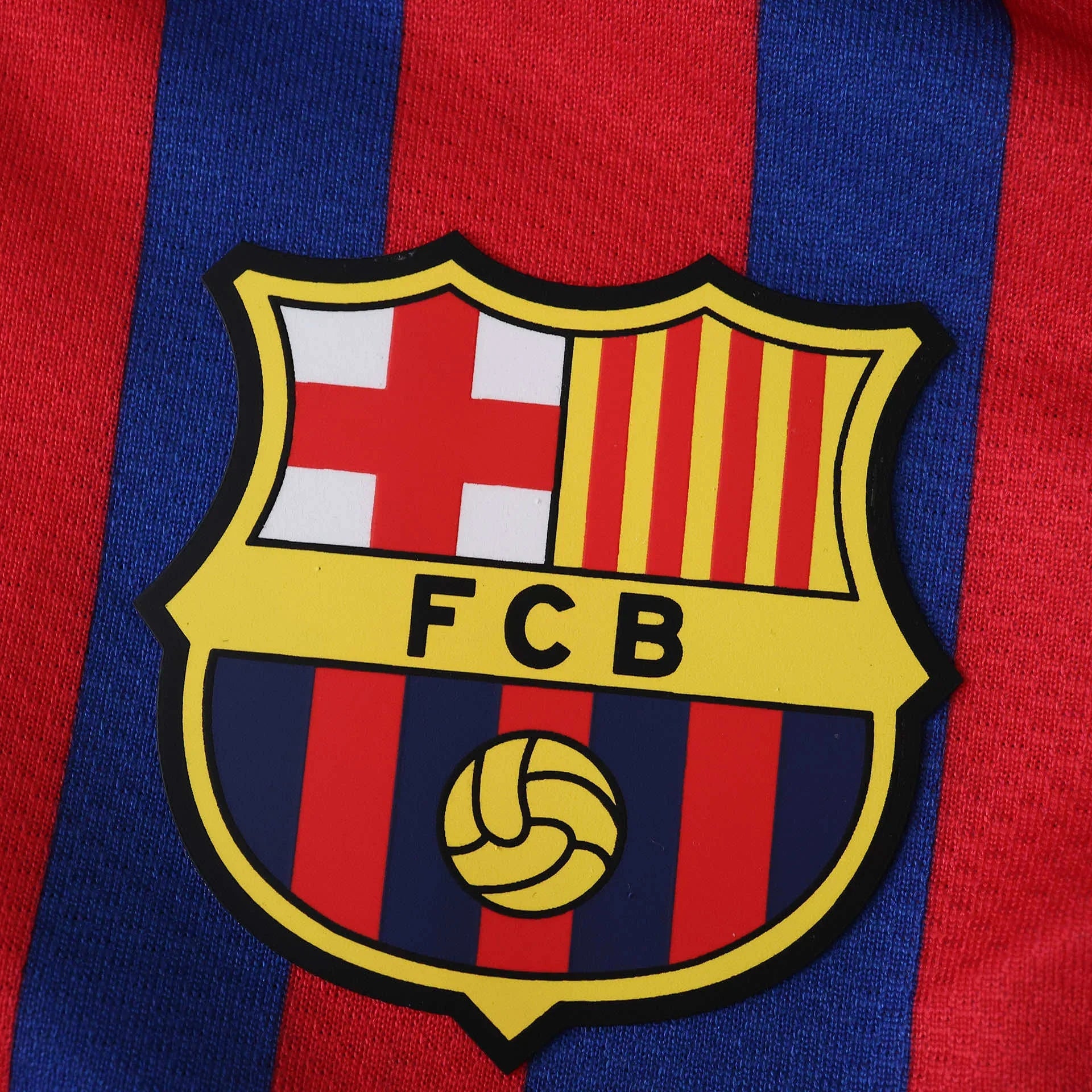 Escudo do FC Barcelona em detalhe, com cores vermelho, azul e amarelo, sobre camisa listrada em vermelho e azul.