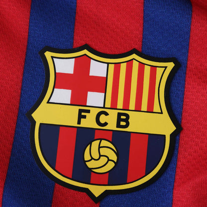 Escudo do FC Barcelona em detalhe, com cores vermelho, azul e amarelo, sobre camisa listrada em vermelho e azul.