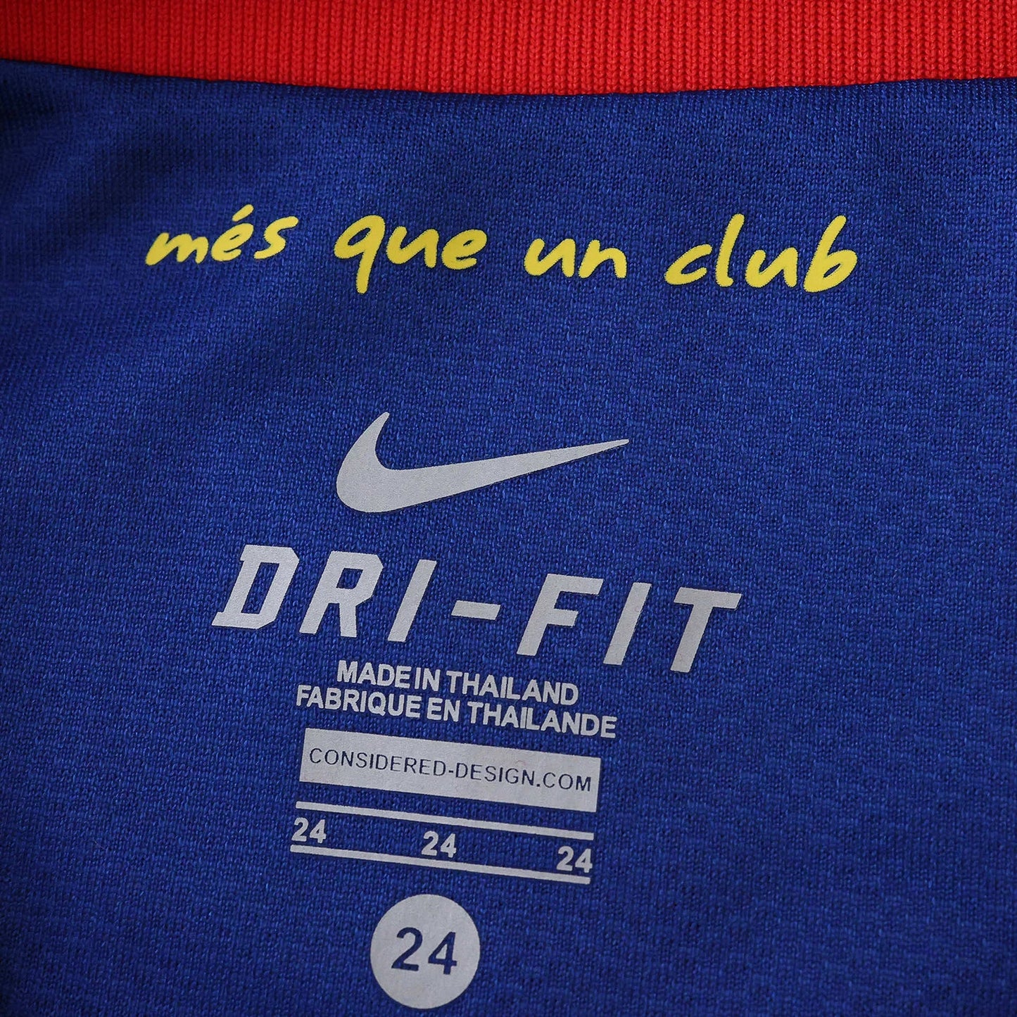 Detalhe da etiqueta interna de uma camisa azul Nike DRI-FIT. Contém o lema 'més que un club' em amarelo, logo Nike e informações de fabricação e tamanho 24.