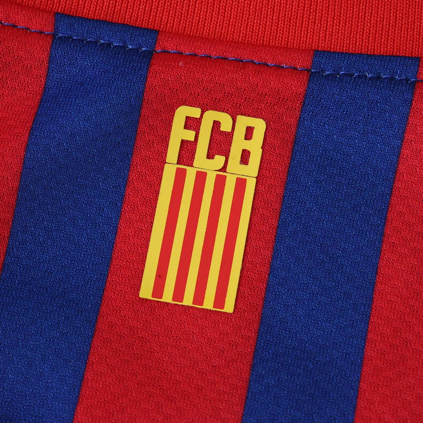 Foto detalhada de camisa de futebol com listras verticais azuis e vermelhas, e logo amarelo 'FCB' com listras vermelhas e amarelas.
