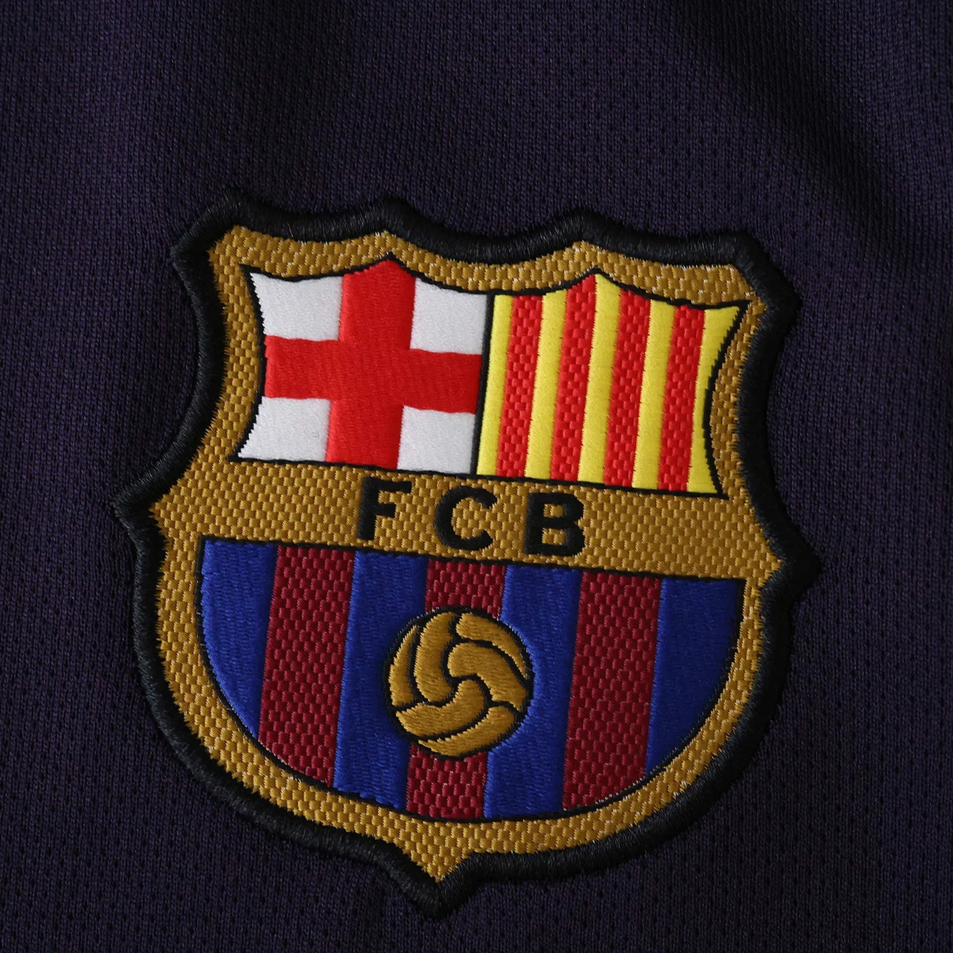 Escudo bordado do FC Barcelona em tecido de camisa azul-marinho. Detalhes em vermelho, amarelo, branco, azul e dourado. Letras FCB e bola.