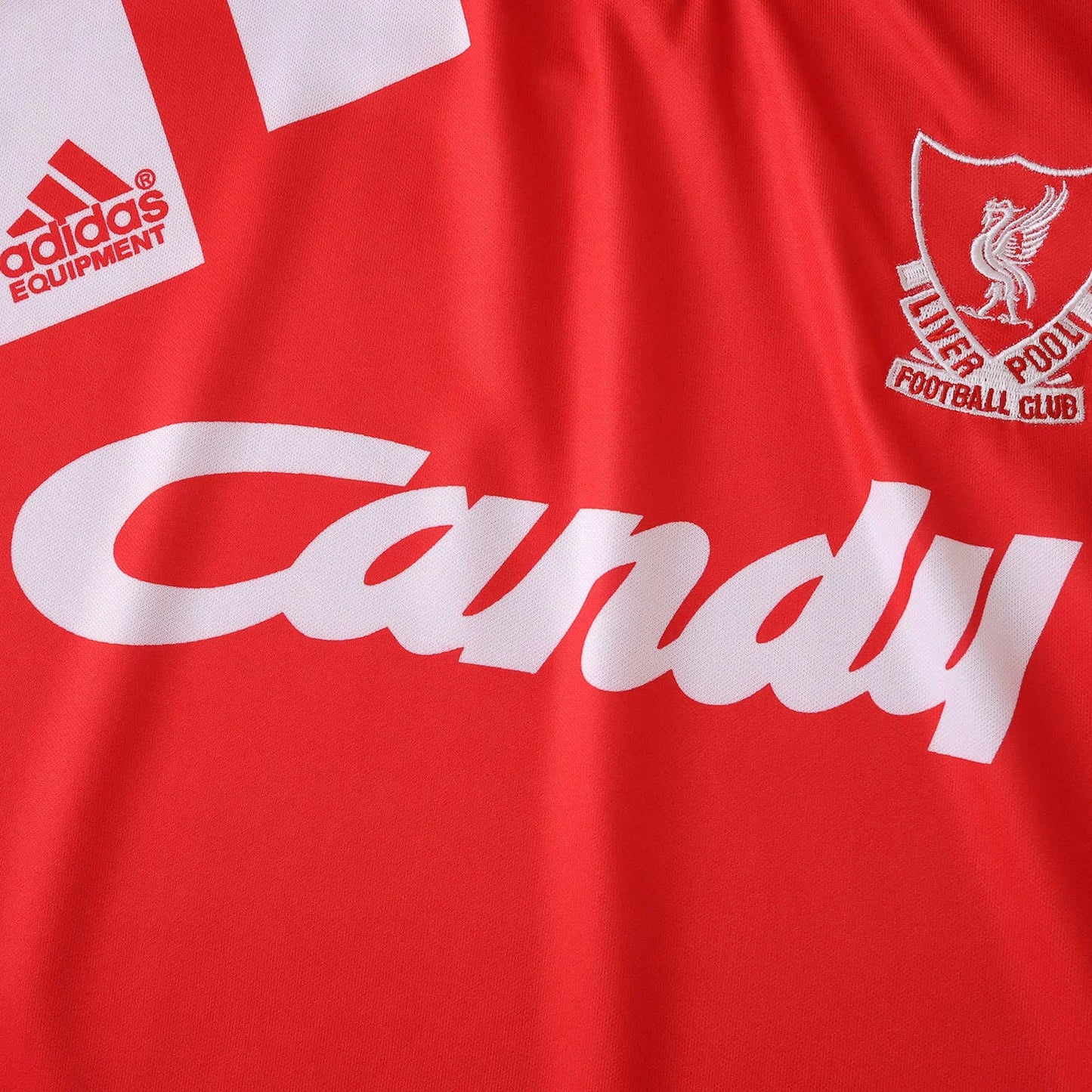 Close-up da camisa de futebol infantil do Liverpool FC, vermelha, com escudo do clube, logo Adidas Equipment e patrocínio Candy em branco.