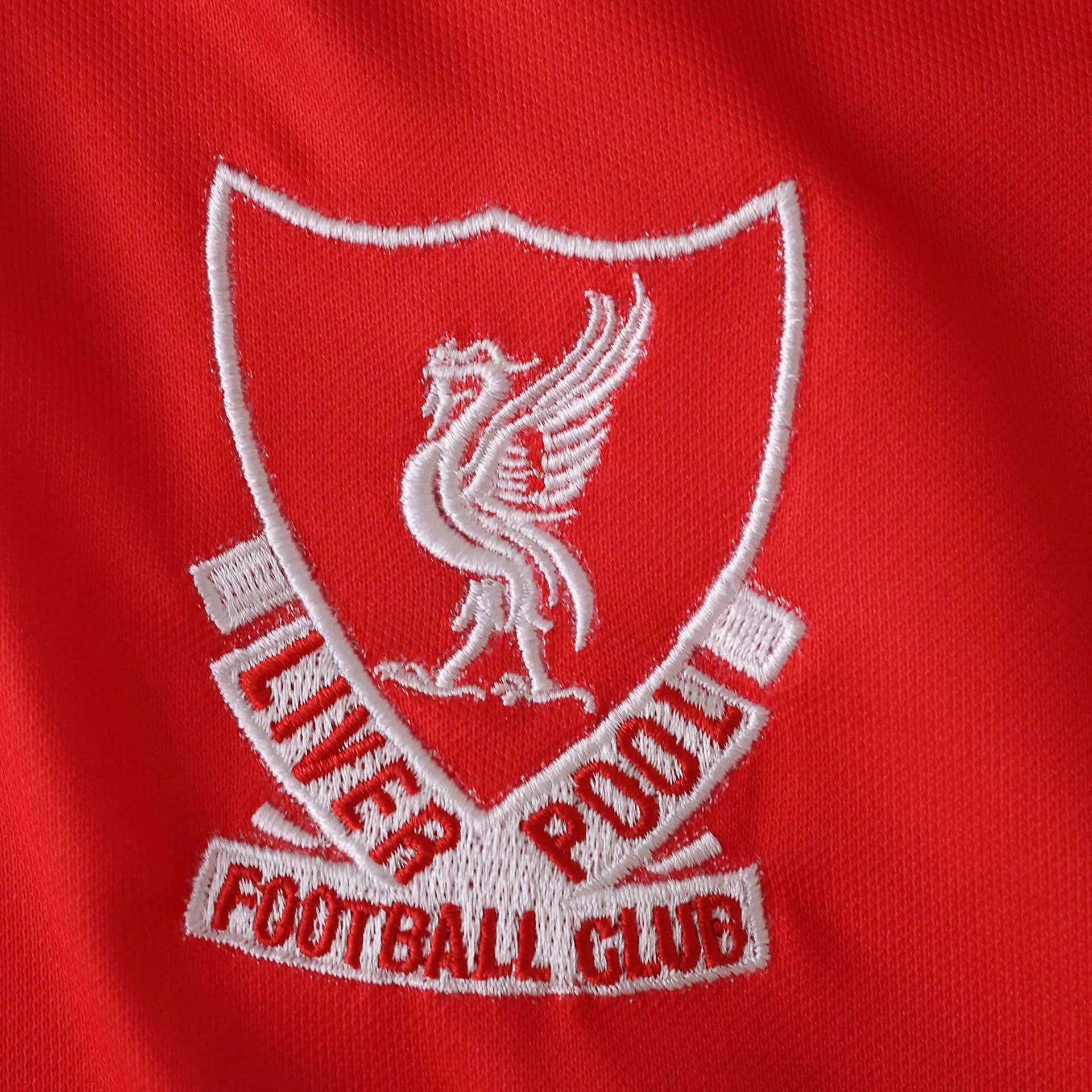 Emblema bordado branco do Liverpool FC em tecido vermelho, mostrando o pássaro Liver e texto 'Liverpool Football Club' em vermelho.