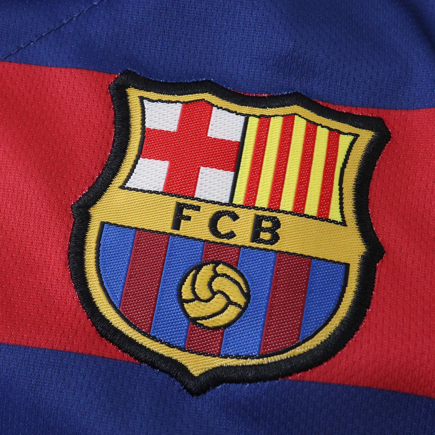 Close-up do escudo bordado do FC Barcelona em uma camisa infantil azul e vermelha. Detalhe da cruz, listras verticais e as letras FCB.