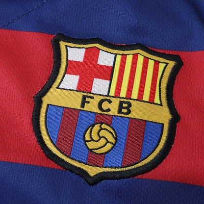 Close-up do escudo bordado do FC Barcelona em uma camisa infantil azul e vermelha. Detalhe da cruz, listras verticais e as letras FCB.