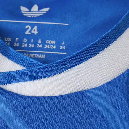 Detalhe de camisa infantil azul com gola listrada branca e azul. Logo Adidas e etiqueta de tamanho 24 visíveis, fabricada no Vietnã.