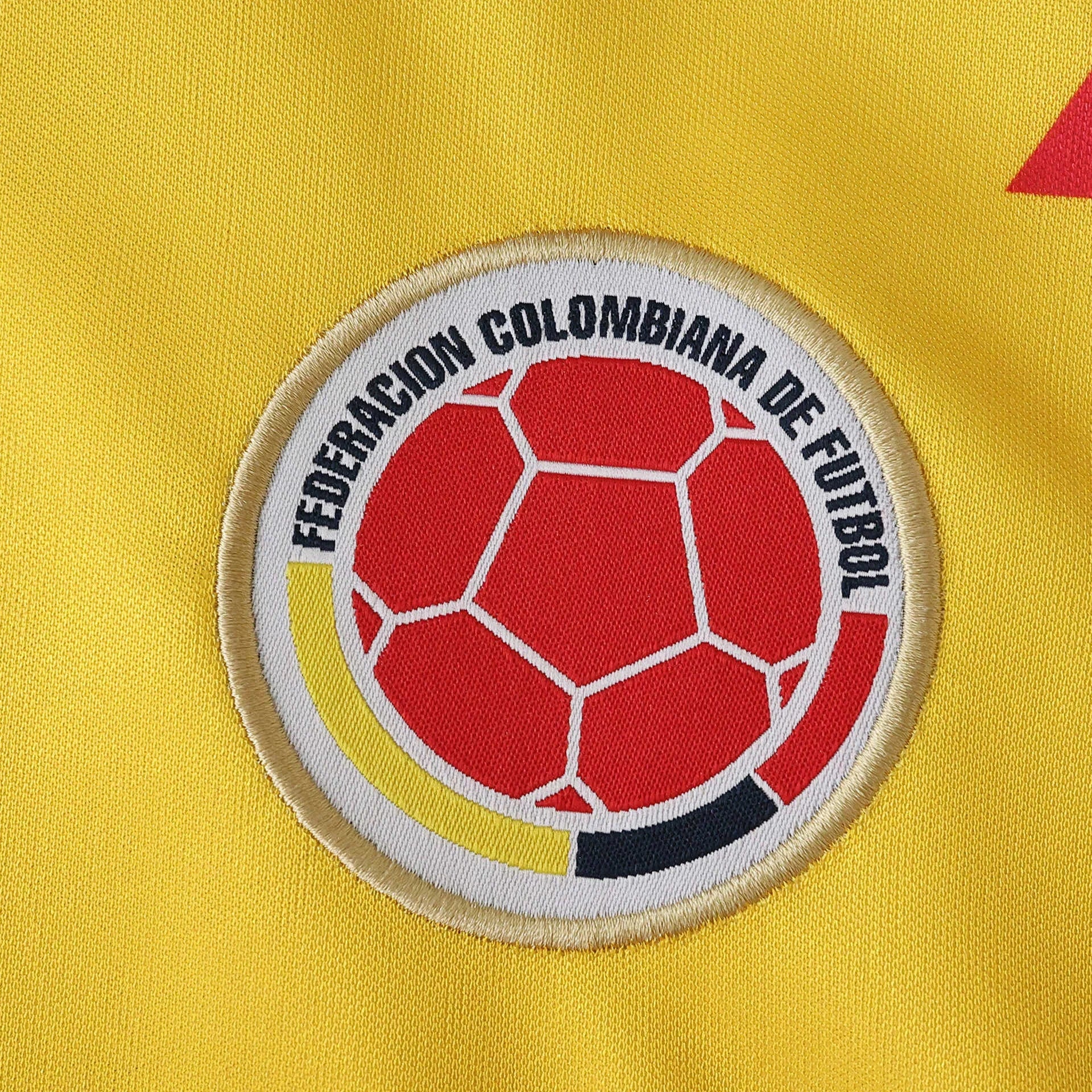 Close-up do escudo da Federação Colombiana de Futebol bordado em camisa de futebol amarela. Bola vermelha e listras amarela, branca, preta.
