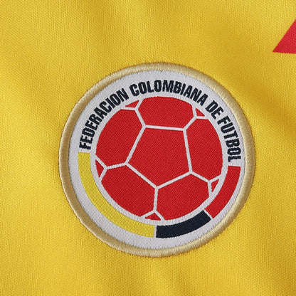 Close-up do escudo da Federação Colombiana de Futebol bordado em camisa de futebol amarela. Bola vermelha e listras amarela, branca, preta.