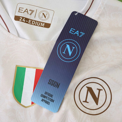 Detalhe de camisa branca do Napoli com logotipos EA7 e N, etiqueta 24-EDIUM, patch de campeão italiano e hang tag oficial.