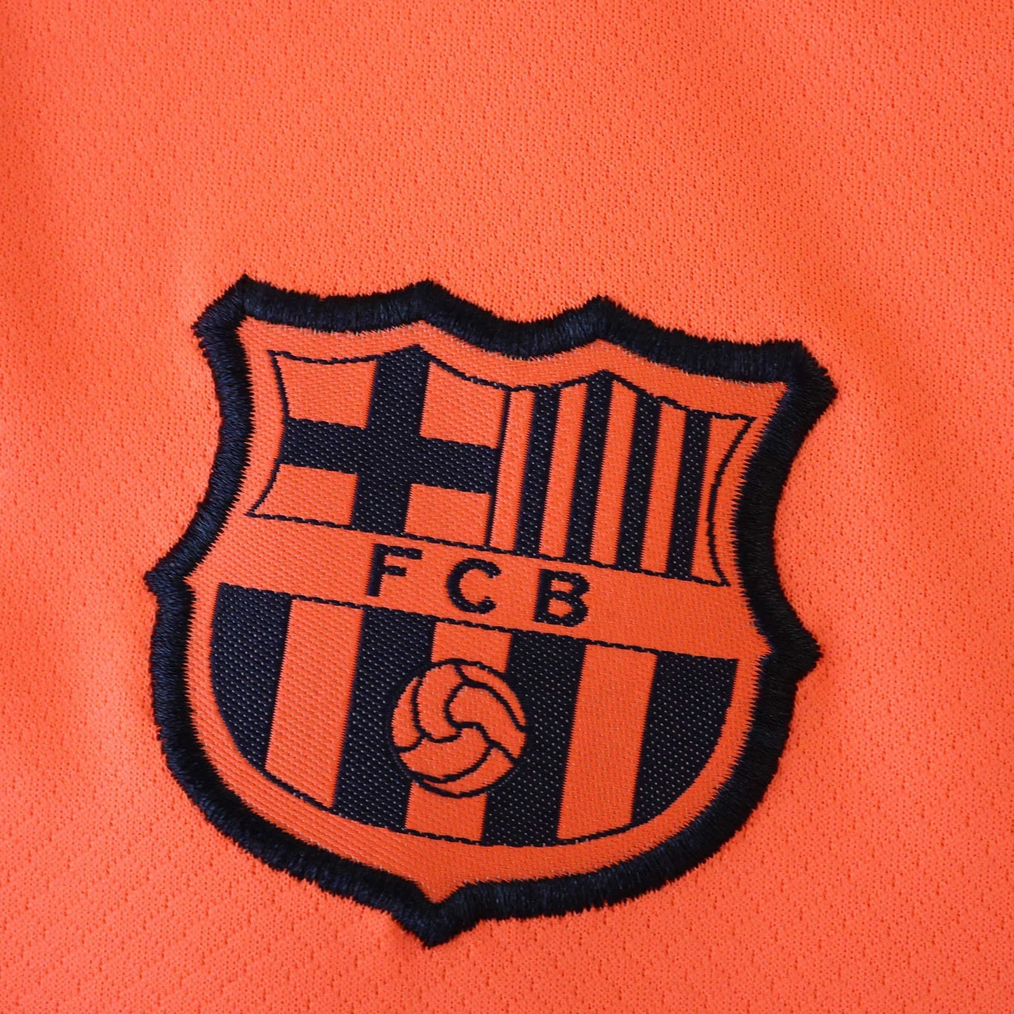 Detalhe do escudo bordado na camisa infantil oficial do FC Barcelona. Parte do kit Mini Mantos laranja
